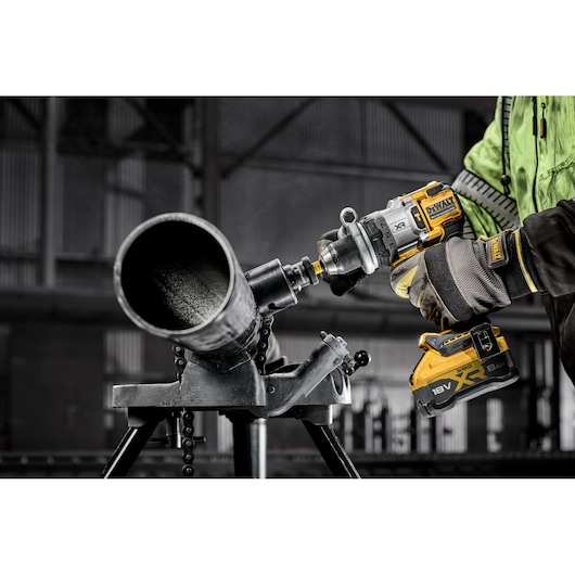 A person wearing gloves and a green jacket uses a DEWALT 18V XR cordless drill to cut into a large metal pipe mounted on a stand in an industrial setting.