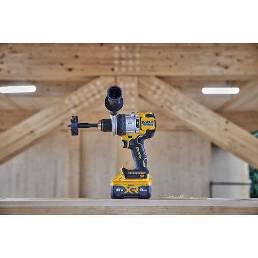 A DEWALT 18V XR cordless drill with a side handle and hole saw attachment is shown standing on a wooden surface in a workshop setting.