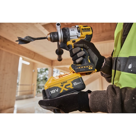 Close-up of a DEWALT cordless drill in use, featuring an 18V XR 8Ah battery. The drill is being held by a person wearing gloves and a high-visibility work vest inside a wooden construction site.