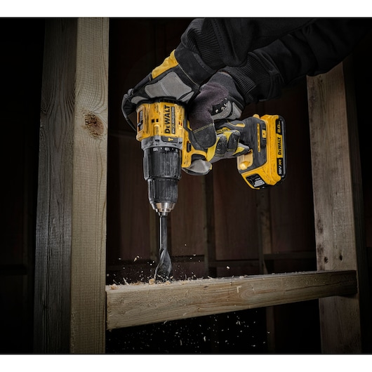 A person wearing work gloves uses a DEWALT cordless drill to drill into a wooden beam, with wood shavings flying from the