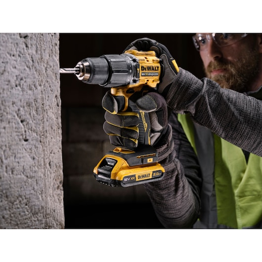 A person wearing work gloves uses a yellow and black DEWALT cordless drill to drill into a concrete wall. The drill is