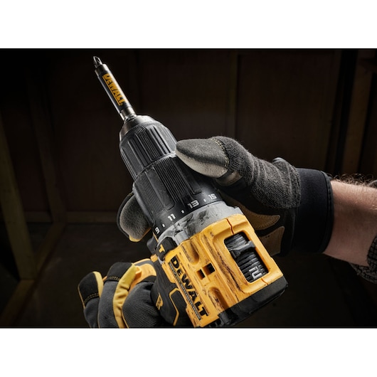 Gloved hand holding a yellow and black DEWALT cordless drill with a screwdriver bit attached.