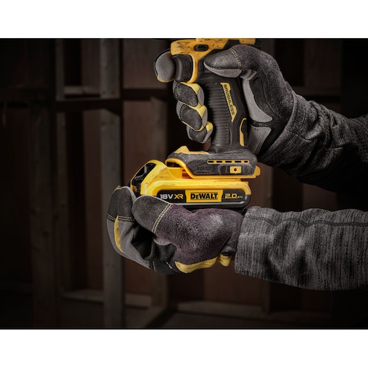 Gloved hands holding a DEWALT 18V XR cordless drill with a 2.0Ah battery in a construction setting.