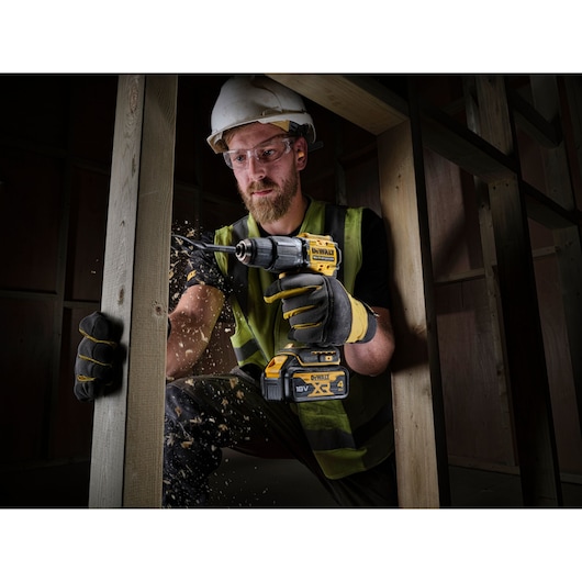 A construction worker wearing safety gloves, a high-visibility vest, and a helmet is using a DEWALT 18V XR cordless drill to