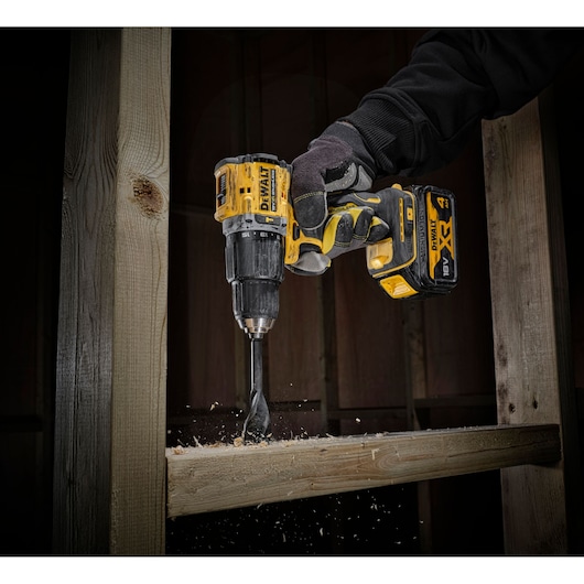 A DEWALT cordless drill being used to bore a hole into a wooden beam, with wood shavings flying, held by a gloved hand.