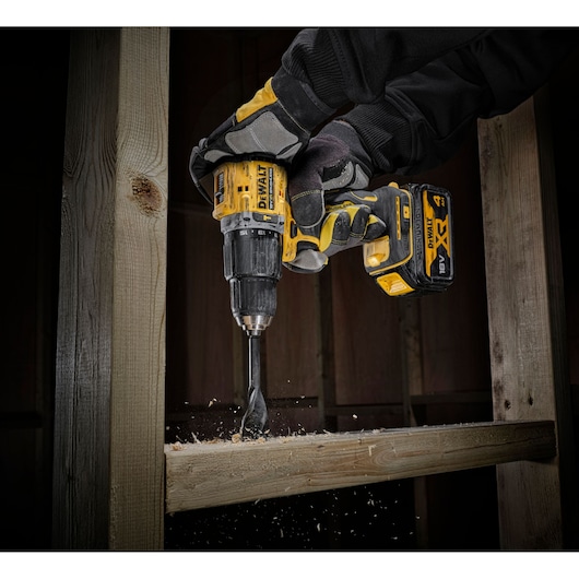 A person wearing protective gloves uses a DEWALT cordless drill to bore a hole in a wooden beam, with wood chips visible