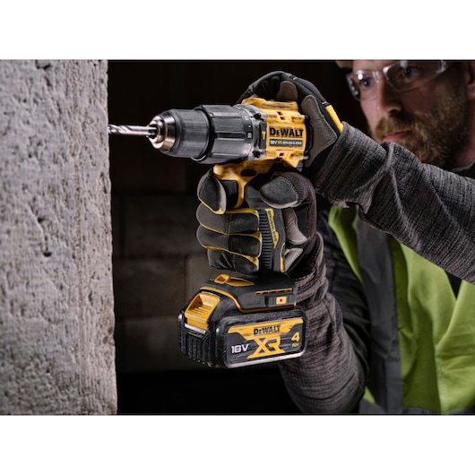 A person wearing work gloves and a safety vest uses a DEWALT cordless drill to drill into a concrete wall. The drill is