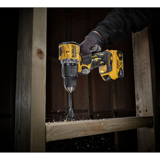 A close-up view of a gloved hand using a yellow DEWALT cordless power drill to bore a hole into a wooden beam, with wood