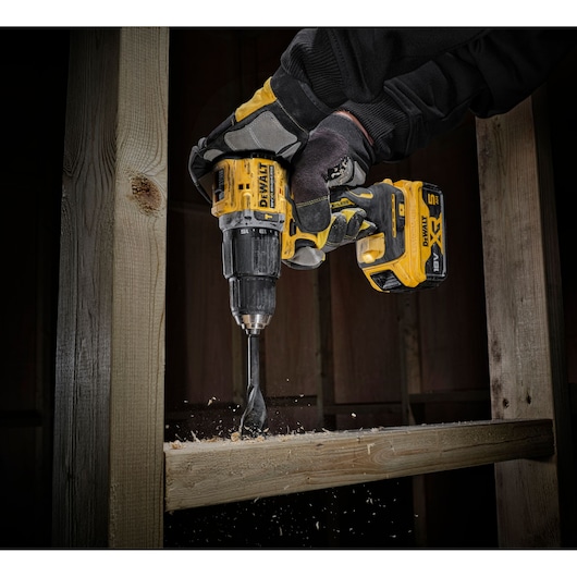 A close-up of a gloved hand using a yellow DEWALT cordless drill to drill into a wooden beam, with wood shavings flying from