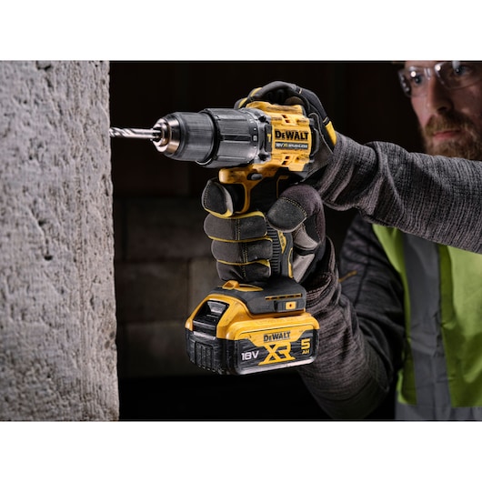 A person wearing gloves is using a DEWALT 18V XR cordless drill to drill into a concrete wall.