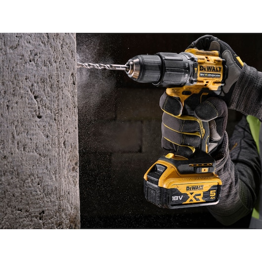 A close-up of a gloved hand holding a DEWALT 18V XR cordless drill with a brushless motor, drilling into a concrete wall.