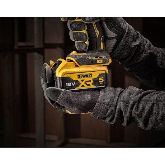 Gloved hands holding a DEWALT cordless tool with an 18V XR 5Ah battery, shown in a workshop setting.
