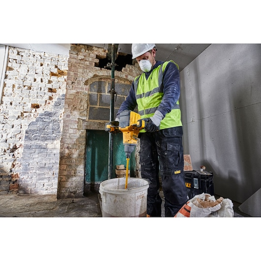 A construction worker wearing a high-visibility vest and hard hat is operating a yellow DEWALT DCD240N paddle mixer to mix materials in a white bucket inside a partially renovated room.
