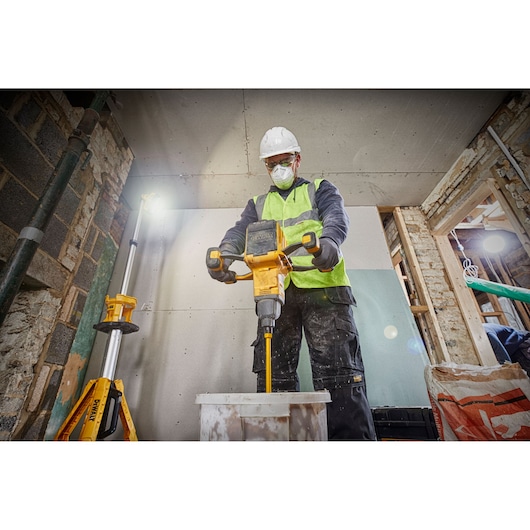 A construction worker wearing a hard hat and high-visibility vest uses a DEWALT DCD240X2 power mixer to mix material in a bucket, inside an unfinished building.