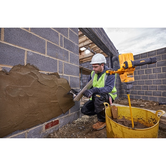 A construction worker wearing a safety helmet and a high-visibility vest applies plaster to a brick wall at a building site. Next to the worker is a yellow DEWALT mixing drill (model DCD240X2) placed in a large yellow container filled with plaster mix.