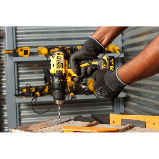 A person wearing work gloves is using a yellow and black DEWALT cordless drill to drill into a piece of metal in a workshop. In the background, several other DEWALT power tools are visible on shelves.