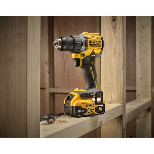 A DEWALT 18V cordless power drill with XR 5 Ah battery is standing upright on a wooden beam at a construction site. Wood shavings and a drill bit are visible nearby.