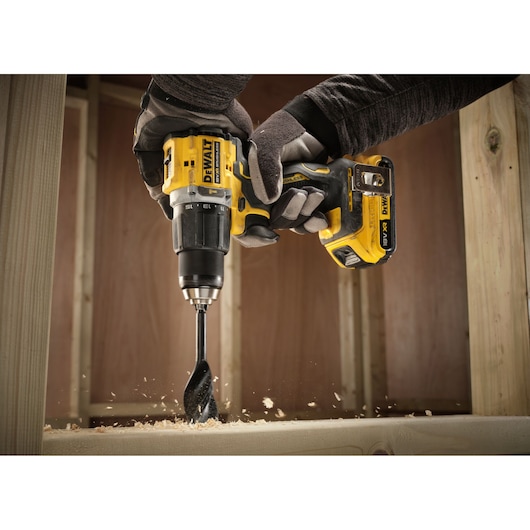 A person wearing protective gloves is using a DEWALT cordless drill with a wood auger bit to drill into a wooden surface