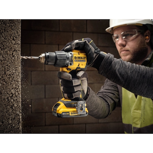 A person wearing gloves is using a DEWALT 18V XR cordless drill to drill into a concrete wall. The drill is yellow and