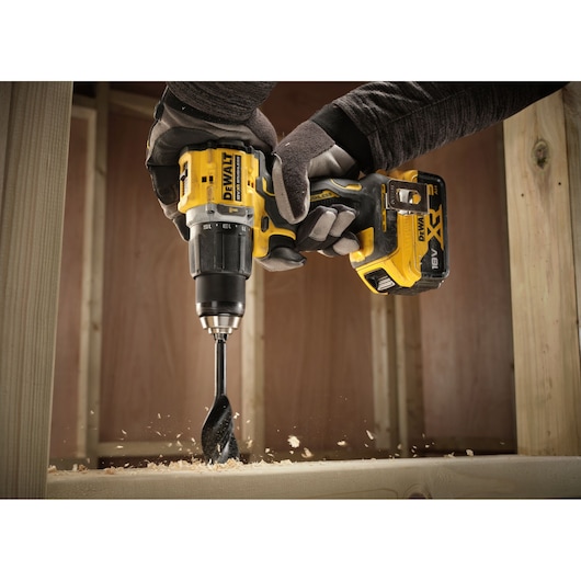 A gloved person using a yellow and black DEWALT cordless drill to drill into a wooden beam, with wood shavings visible