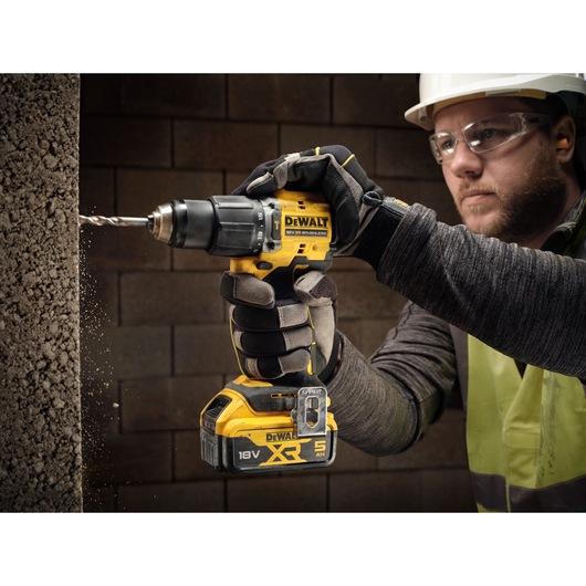 A person wearing protective gloves uses a DEWALT 18V XR brushless cordless drill to drill into a concrete wall. The image