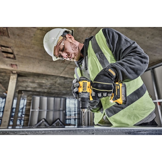 A construction worker wearing a safety helmet, high-visibility vest, and gloves is using a DEWALT DCD800E2T cordless drill to drill into a metal surface at an indoor construction site.