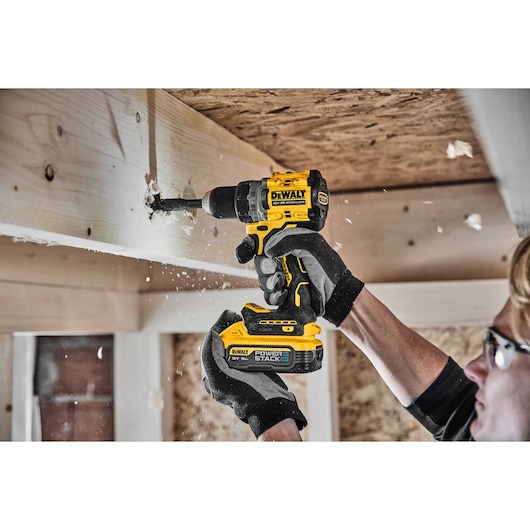 A person wearing gloves uses a DEWALT DCD800H2T cordless drill to make a hole in a wooden beam, with wood shavings flying