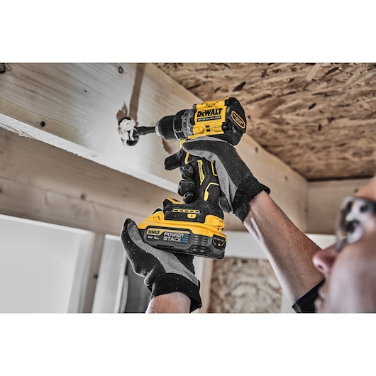 A person wearing gloves is using a DEWALT DCD800H2T cordless drill with a POWERSTACK battery to drill a hole in a wooden