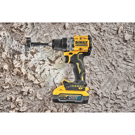 DEWALT cordless drill with PowerStack battery drilling through a textured surface with wood shavings visible.