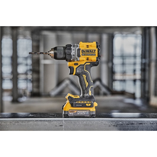 A DEWALT cordless power drill with an 18V POWERSTACK battery is standing upright on a metal surface in an industrial setting. The tool is yellow and black, with a drill bit attached.