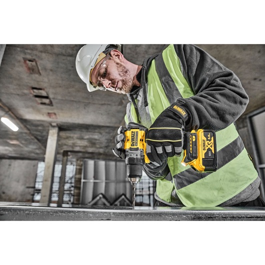 A person wearing safety gear, including gloves, a helmet, and a hi-vis vest, is using a DEWALT DCD800P2T cordless drill to work on a metal surface in an industrial environment.