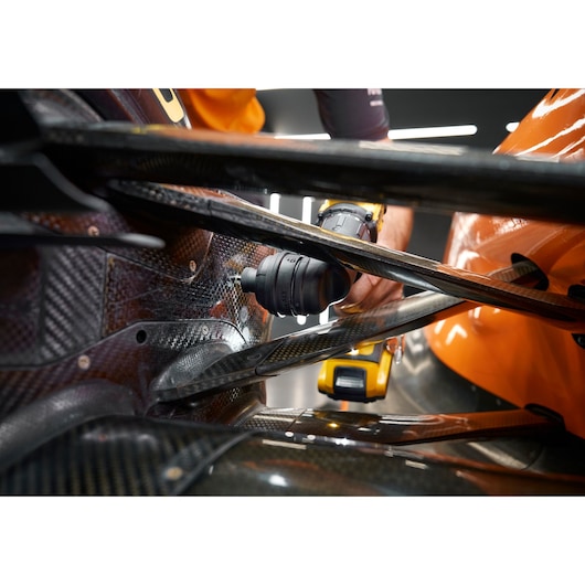 A close-up of a DEWALT cordless drill being used to work on the carbon fiber body of an orange vehicle, likely in an automotive or motorsport setting.