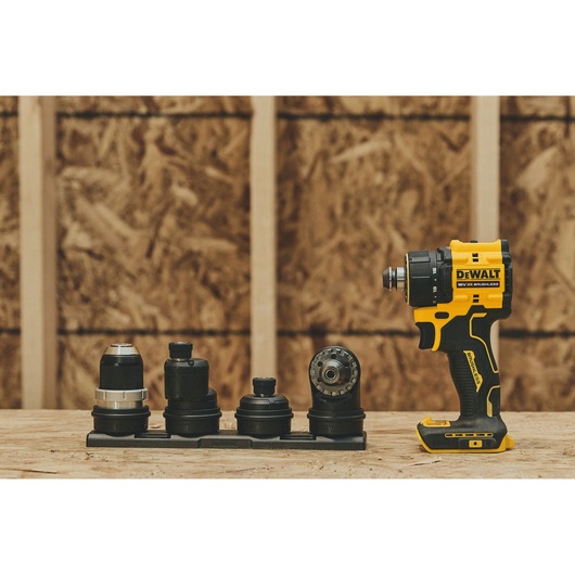A DEWALT cordless drill driver with four interchangeable attachments displayed on a wooden surface, set against an unfinished plywood wall background.