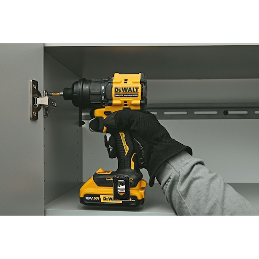 A DEWALT 18V XR brushless cordless drill being used by a gloved hand to install a hinge inside a grey cabinet.