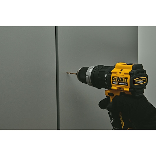 A close-up of a gloved hand using a DEWALT cordless drill with a brushless motor to drill into a gray wall.