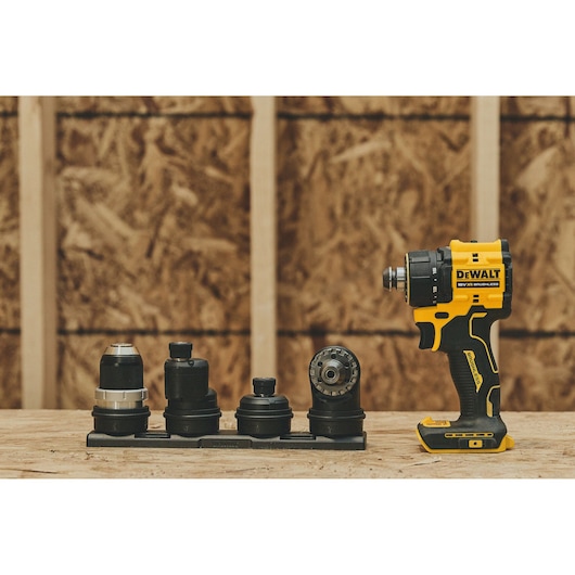 A DEWALT cordless drill with a yellow and black design is placed on a wooden surface. Next to it, there is a set of multiple interchangeable drill attachments organized in a row. The background shows wooden construction panels.