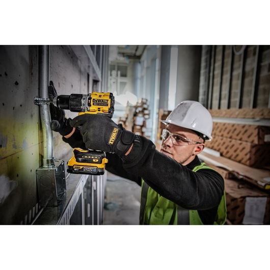 A person wearing a hard hat, safety vest, and gloves uses a DEWALT DCD805E1T power drill to work on a metal conduit attached to a wall in an industrial or construction setting.