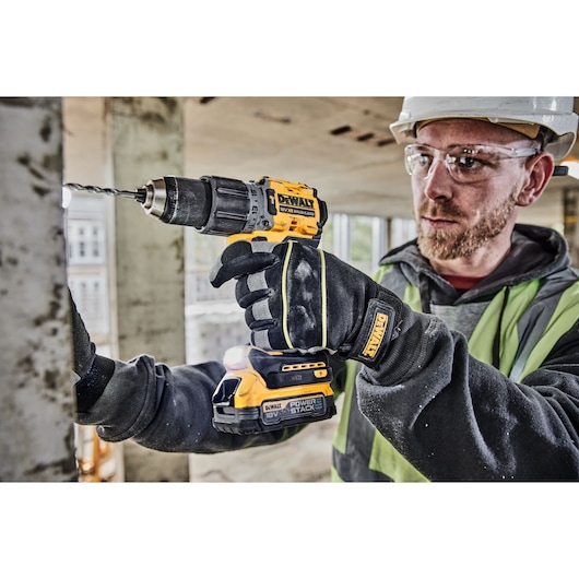 A person wearing work gloves and safety gear is drilling into a metal surface using a yellow and black DEWALT cordless power drill at a construction site.