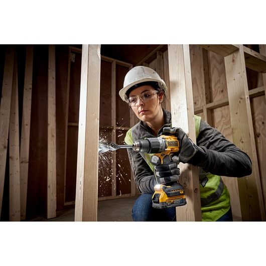 A person wearing a hard hat and safety vest is using a DEWALT cordless drill to bore a hole into a wooden beam inside a