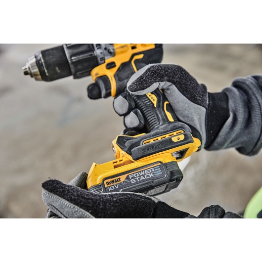 Close-up of a person wearing gloves holding a DEWALT cordless drill with a POWERSTACK 18V battery attached.