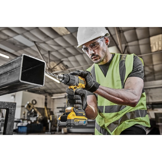 A person wearing a high-visibility vest and hard hat is using a DEWALT cordless drill to drill into a metal square tube in