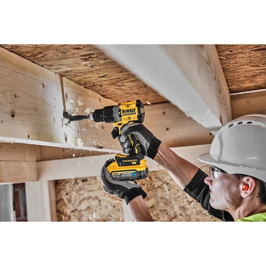 A person wearing gloves and a safety helmet is using a DEWALT cordless drill to make a hole in a wooden beam. Wood shavings
