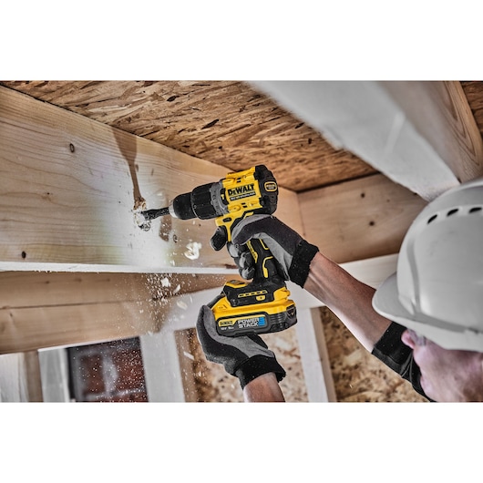 A person wearing gloves and a helmet uses a DEWALT cordless drill to drill into a wooden beam, with wood shavings flying.