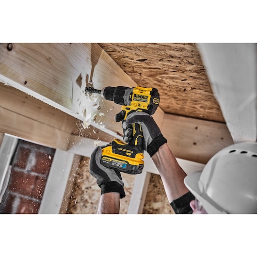 A person wearing safety gloves and a helmet uses a yellow DEWALT cordless drill to bore into a wooden beam, causing wood