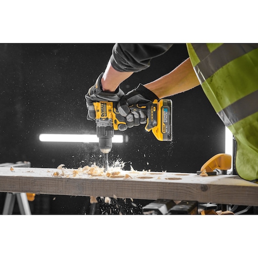 A person wearing gloves and a high-visibility vest uses a DEWALT cordless drill to drill into a wooden plank, creating wood