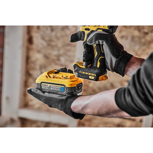 Gloved hands holding a DEWALT PowerStack 18V battery and cordless drill in a workshop setting.