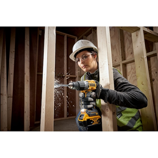 A person wearing safety gear is using a DEWALT DCD805 cordless drill to drill into a wooden beam at a construction site. Sawdust is visible as the drill bit penetrates the wood.