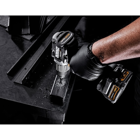 A close-up of a person wearing a black glove using a DEWALT cordless drill to drill into a metal rectangular tube. Metal shavings are visible on the surface.