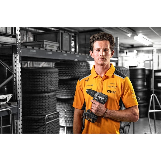 A person wearing an orange and grey shirt is standing in an industrial storage area with stacks of tires and holding a DEWALT cordless drill (model DCD85ME2GT). The face is blurred for privacy.