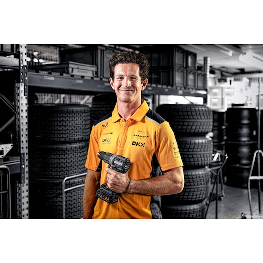 A person with a blurred face wearing an orange and black McLaren team shirt is holding a DEWALT cordless drill (model DCD85ME2GT) in a workshop with stacked tires and shelving in the background.